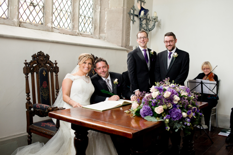 A bride and groom, sitting and smiling as they sign the register